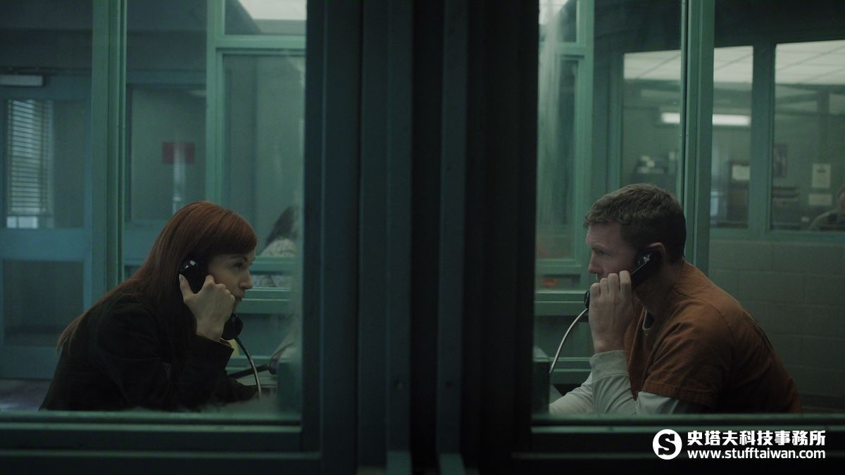 "A woman and a man in prison uniform sit across glass in a visitation room using phones to talk. The setting is dimly lit with institutional green walls and other people are visible in the background."